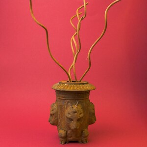 Peut inclure: Un vase décoratif de couleur bronze vieilli avec des détails de têtes et de pattes de lion. Le vase contient plusieurs branches sèches et torsadées. Le vase a une base ronde et un large bord décoratif. Le fond est rouge uni.