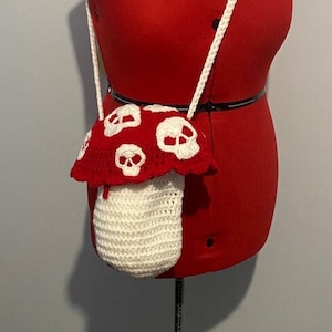 May include: A white crocheted mushroom-shaped bag with a red crocheted top featuring white skull designs. The bag has a white crocheted strap.