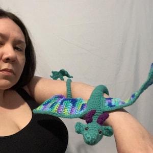 May include: A green crocheted dragon with purple accents wrapped around a person's arm. The dragon has a long tail and wings.