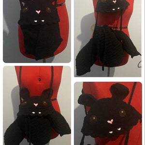 May include: A black crocheted bat-shaped bag with a cute face and wings. The bag has a pink nose and white teeth. The bag is hanging on a red mannequin.