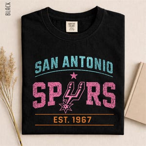 May include: A black crew neck t-shirt featuring a distressed graphic print. The text reads SAN ANTONIO in light blue, SPURS in pink with a pink team logo, and EST. 1967 in orange. The shirt has a Comfort Colors brand label inside the neckline.