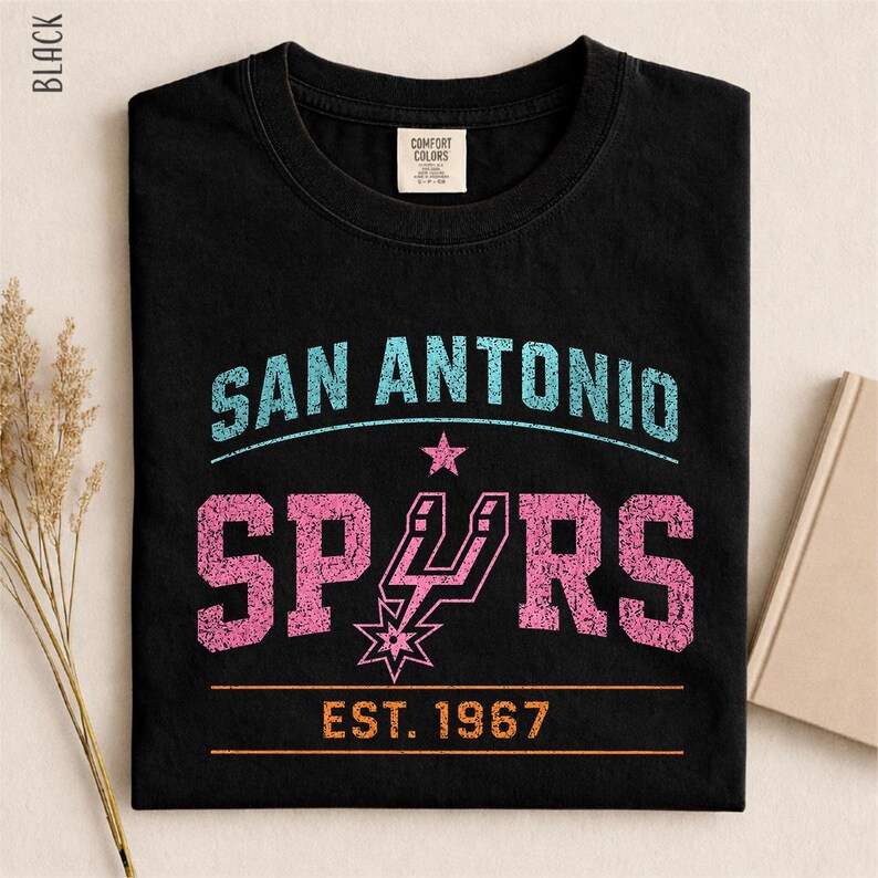 May include: A black crew neck t-shirt featuring a distressed graphic print. The text reads SAN ANTONIO in light blue, SPURS in pink with a pink team logo, and EST. 1967 in orange. The shirt has a Comfort Colors brand label inside the neckline.
