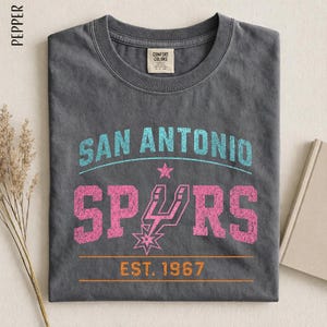 May include: A charcoal gray t-shirt featuring the text SAN ANTONIO in light blue, SPURS in pink with a star-shaped logo, and EST. 1967 in orange. The shirt has a crew neckline and a Comfort Colors brand label on the inside of the collar.