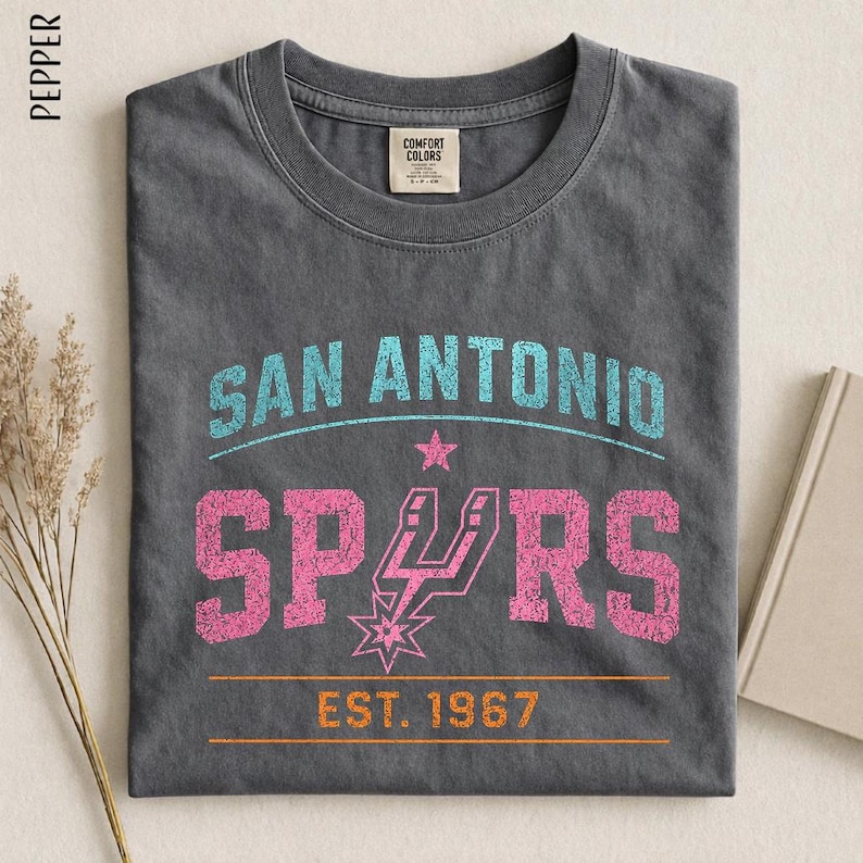 May include: A charcoal gray t-shirt featuring the text SAN ANTONIO in light blue, SPURS in pink with a star-shaped logo, and EST. 1967 in orange. The shirt has a crew neckline and a Comfort Colors brand label on the inside of the collar.