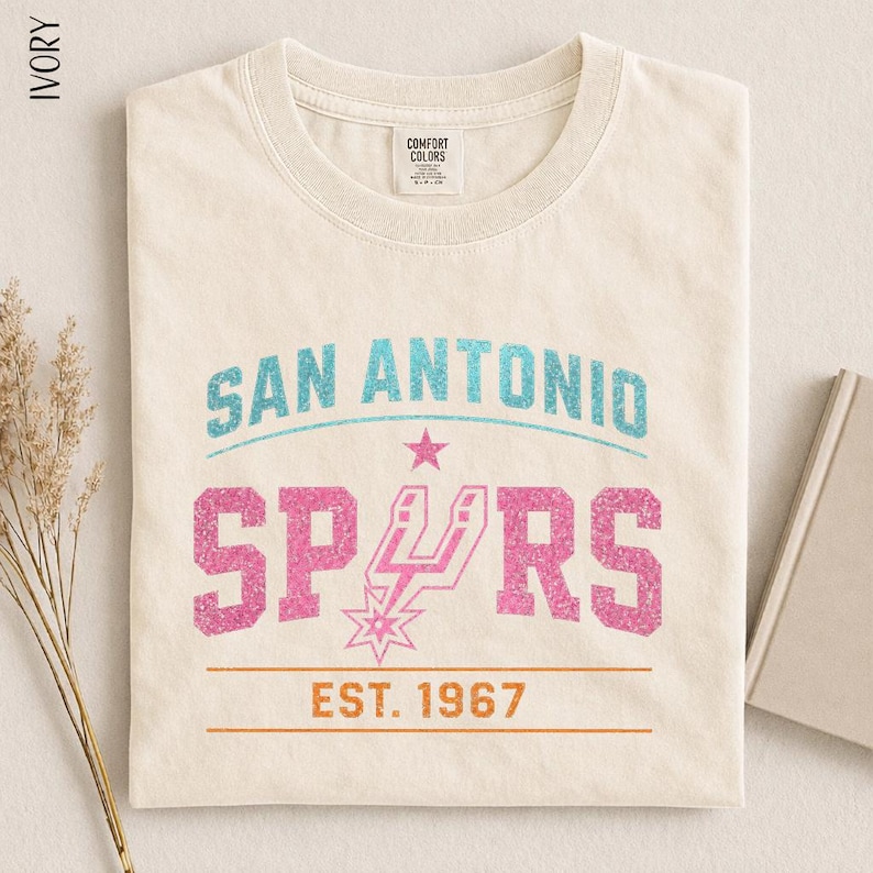 May include: An ivory crew neck t-shirt featuring a graphic print with the text SAN ANTONIO in teal, SPURS in pink with a pink star logo, and EST. 1967 in orange between two horizontal lines. The shirt has a Comfort Colors brand label at the neck.