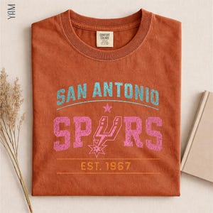 May include: A rust-colored crew neck t-shirt featuring a distressed graphic print. The text reads SAN ANTONIO in light blue, SPURS in pink with a team logo replacing the U, and EST. 1967 in yellow. The shirt has a Comfort Colors brand neck label.
