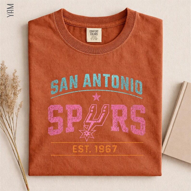 May include: A rust-colored crew neck t-shirt featuring a distressed graphic print. The text reads SAN ANTONIO in light blue, SPURS in pink with a team logo replacing the U, and EST. 1967 in yellow. The shirt has a Comfort Colors brand neck label.