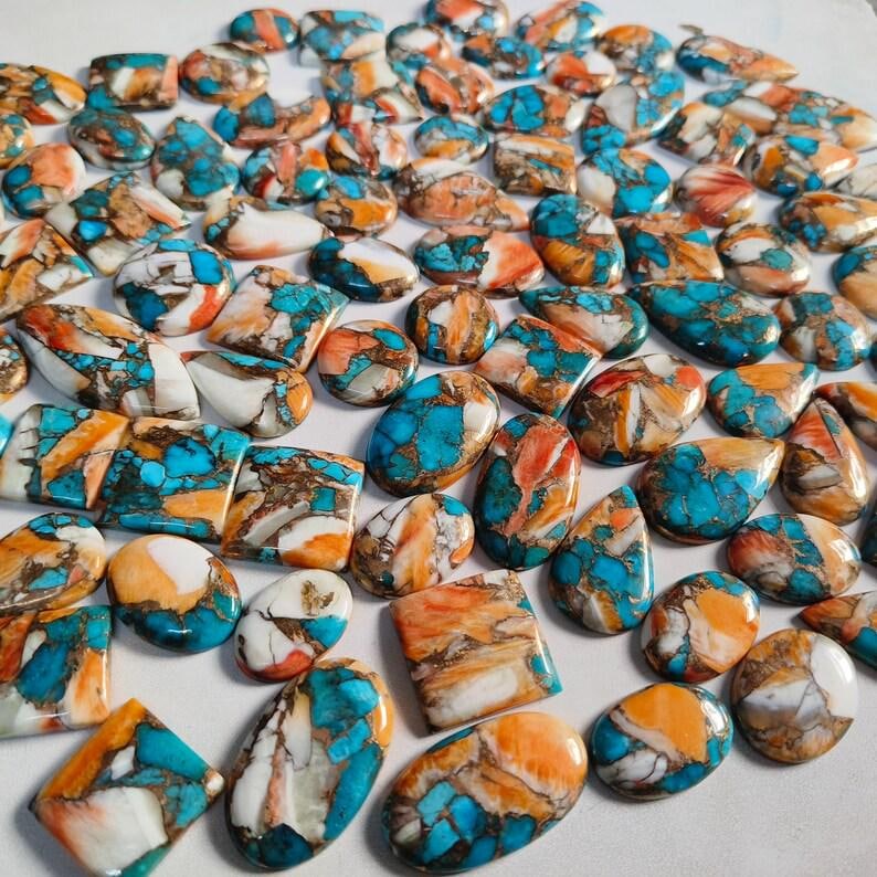 May include: A collection of polished turquoise, orange, and white gemstone cabochons in various shapes. The gemstones have a marbled appearance with a mix of colors and patterns. The cabochons are oval, rectangular, and teardrop-shaped.