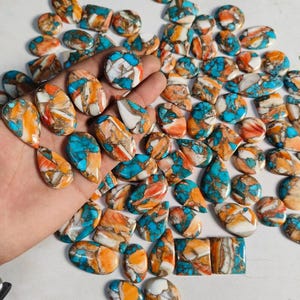 May include: A collection of polished gemstones in various shapes, including ovals, teardrops, and squares. The stones feature a mix of turquoise, orange, and white colors, with a marbled appearance. The gemstones are displayed on a white surface.