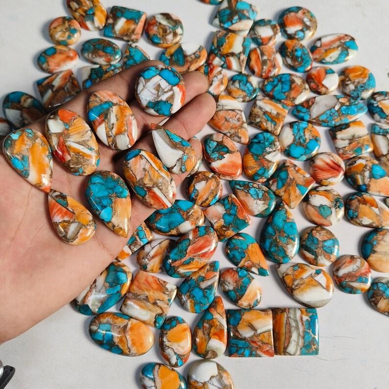May include: A collection of polished gemstones in various shapes, including ovals, teardrops, and squares. The stones feature a mix of turquoise, orange, and white colors, with a marbled appearance. The gemstones are displayed on a white surface.