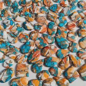 May include: A collection of polished gemstones in various shapes, including ovals, teardrops, and squares. The stones feature a mix of turquoise, orange, brown, and white colors, with a marbled or speckled appearance. The gemstones are arranged closely together.