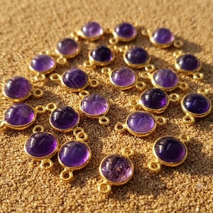 Puede incluir: Una colección de dijes redondos de amatista morada engastados en biseles de metal dorado. Cada dije presenta una superficie lisa y pulida, con dos pequeñas anillas circulares en los lados opuestos para conectar con piezas de joyería.