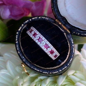 May include: A rectangular ring with alternating rows of small, round, clear stones and square, red stones. The ring is set in a black velvet jewelry box, surrounded by white and purple flowers.