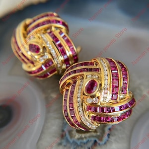 May include: A pair of gold-toned earrings with a knot design. The earrings are adorned with square-cut rubies and small diamonds. Each earring features an oval ruby in the center. The jewelry is displayed on a light-colored surface.