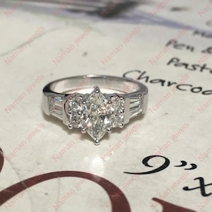 May include: A silver ring with a marquise-cut center stone and smaller stones on either side. The ring has a classic design with a polished finish. The ring is set on a white surface with text that reads "Naman jewels".