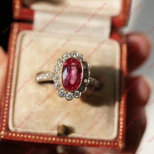 May include: A vintage-style ring with a large, oval-cut ruby center stone. The ruby is surrounded by a halo of small, round diamonds and set in a gold and silver band. The ring is displayed in a red jewelry box.