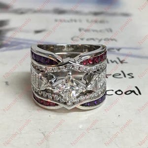 Puede incluir: Un anillo ancho, plateado, con un diamante central de corte cuadrado, rodeado de diamantes más pequeños y filas alternas de piedras preciosas rojas y moradas. El anillo tiene un diseño intrincado y en capas.