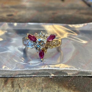 May include: A gold ring with a central round diamond, surrounded by smaller diamonds and three ruby-colored marquise-cut stones. The ring has a band with additional small diamonds.