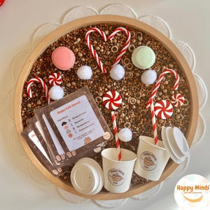 May include: A round wooden sensory bin filled with brown filler, featuring two white paper cups with Happy Minds Cafe logos, red and white striped straws, candy canes, peppermint swirls, white pom-poms, and a Happy Cafe Special menu card.