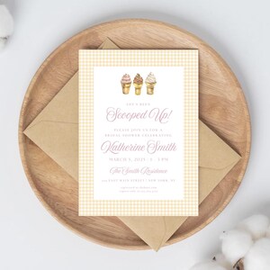 May include: A bridal shower invitation with a light yellow gingham pattern border and a white center. The invitation features watercolor ice cream cones and the text "Scooped Up!" in a script font. The invitation is placed on a wooden tray with a brown envelope.