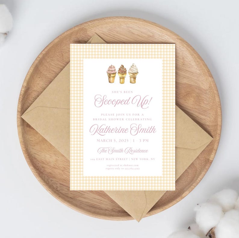 May include: A bridal shower invitation with a light yellow gingham pattern border and a white center. The invitation features watercolor ice cream cones and the text "Scooped Up!" in a script font. The invitation is placed on a wooden tray with a brown envelope.
