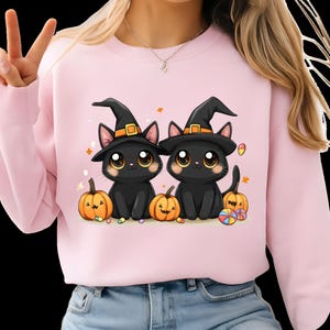 Könnte beinhalten: Rosa Sweatshirt mit einem Halloween-Motiv. Das Design zeigt zwei schwarze Katzen mit Hexenhüten, umgeben von Kürbissen und Süßigkeiten. Die Katzen haben große Augen und sitzen nebeneinander.