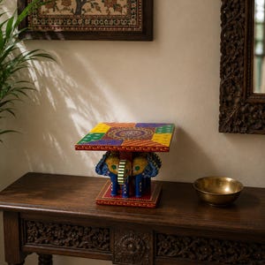 Pode incluir: Uma pequena mesa colorida com um design de elefante. A mesa tem um tampo quadrado com um padrão de patchwork em amarelo, verde, vermelho e roxo. A base apresenta duas figuras de elefante azuis. A mesa está sobre uma superfície de madeira escura.
