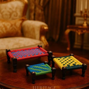 May include: Three miniature woven beds in red, blue, and yellow with dark wooden legs. The beds are displayed on a dark wooden table. The text "HERITAGE CREATIONS" is visible on the table.