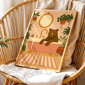 以下が含まれることがあります： ピンクのバスタブでくつろぐ茶色のクマが描かれた、額入りのアートプリント。イラストには、金色のフレームの鏡、植物、ストライプのラグが含まれています。このアートワークは、暖色系の色使いと気まぐれなテーマを持っています。