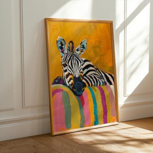 Op de afbeelding: Een ingelijste kunstdruk met een zebra met zwart-witte strepen, rustend op een kleurrijk gestreept kussen. De achtergrond is een levendig geel en de lijst is lichtbruin.
