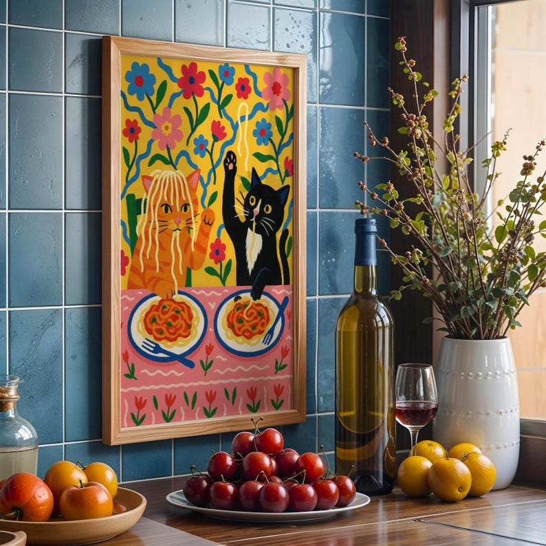 May include: A framed art print featuring two cartoon cats enjoying spaghetti. One cat is orange, the other is black. The background is yellow with colorful flowers. The print is in a light wood frame, displayed on a blue tiled wall.