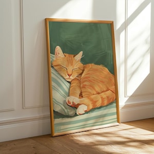 May include: A framed art print featuring a sleeping orange tabby cat. The cat is curled up on a striped pillow against a green background. The artwork is in a wooden frame and leans against a white wall.
