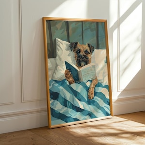 Può includere: Stampa incorniciata raffigurante un cane a letto che legge un libro. Il cane è sotto una coperta a righe blu e bianche, appoggiato su un cuscino bianco. L'opera d'arte ha una cornice in legno ed è appoggiata a una parete bianca e un pavimento in legno.
