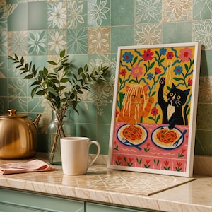 May include: A framed art print featuring two cartoon cats dining on spaghetti. One cat is orange, the other is black. The background is yellow with floral designs. The print is displayed on a countertop with a mug and a teapot.