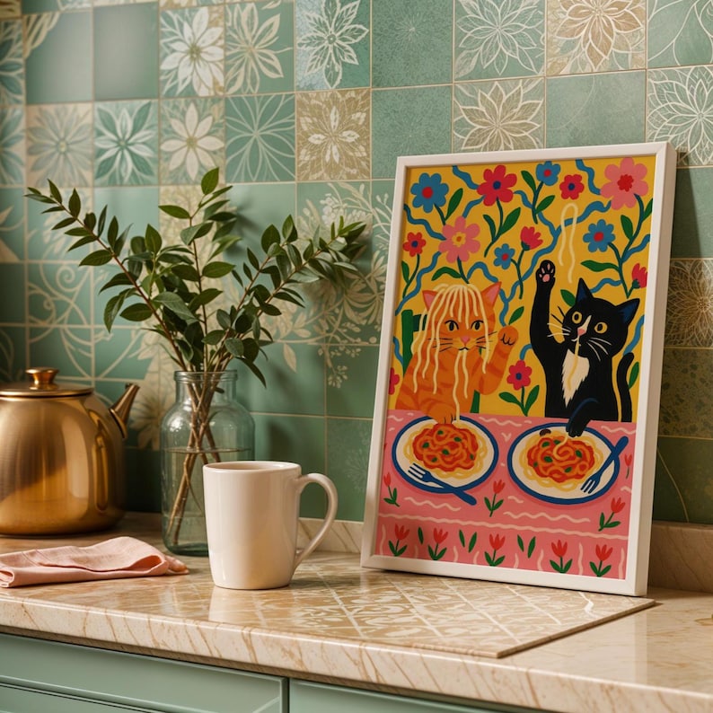 May include: A framed art print featuring two cartoon cats dining on spaghetti. One cat is orange, the other is black. The background is yellow with floral designs. The print is displayed on a countertop with a mug and a teapot.