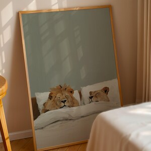 May include: A framed print depicts two lions resting in a bed. The artwork shows a male lion and a lioness lying on pillows under a white duvet. The frame is made of light-colored wood.