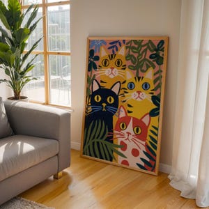 May include: A framed art print featuring a colorful illustration of four stylized cats. The cats have yellow, black, and red fur, with large eyes and surrounded by green and blue foliage. The artwork is displayed in a room with natural light.