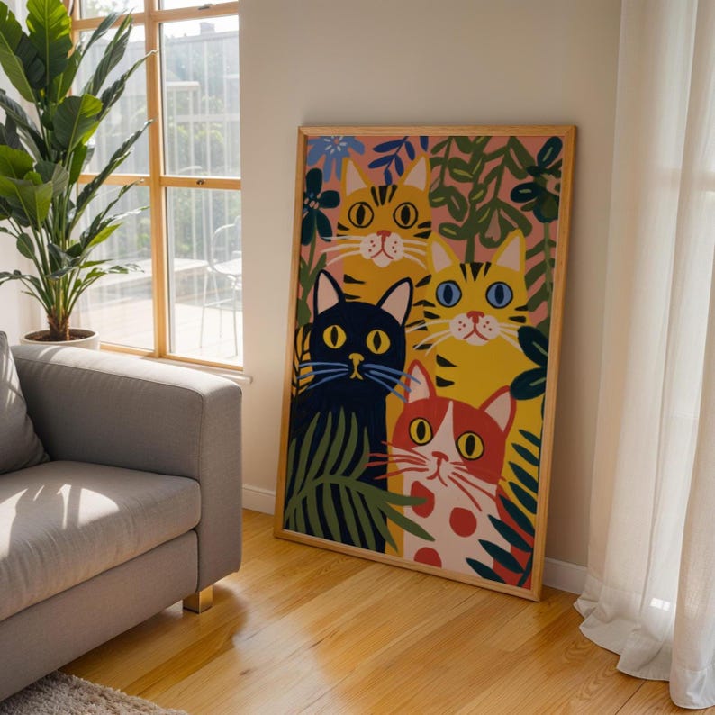 May include: A framed art print featuring a colorful illustration of four stylized cats. The cats have yellow, black, and red fur, with large eyes and surrounded by green and blue foliage. The artwork is displayed in a room with natural light.