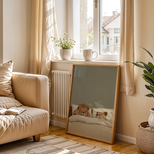 May include: A framed print featuring a lion and lioness in bed, resting under a blanket. The artwork has a light wood frame and is displayed in a room with a beige sofa, a potted plant, and a window with sheer curtains.