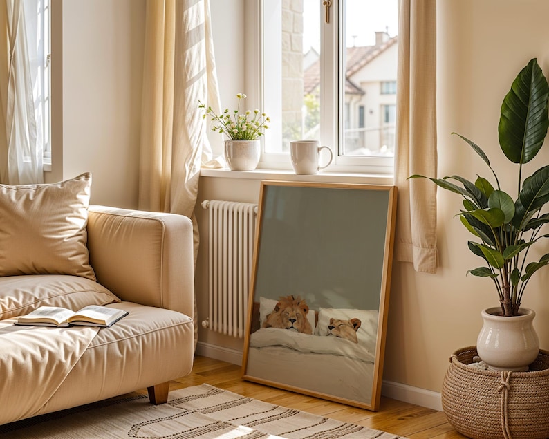 May include: A framed print featuring a lion and lioness in bed, resting under a blanket. The artwork has a light wood frame and is displayed in a room with a beige sofa, a potted plant, and a window with sheer curtains.