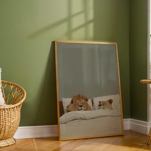 May include: A framed art print featuring a whimsical illustration of two lions in bed, resting on white pillows and under a white blanket. The artwork is set in a room with a green wall and a wooden floor.