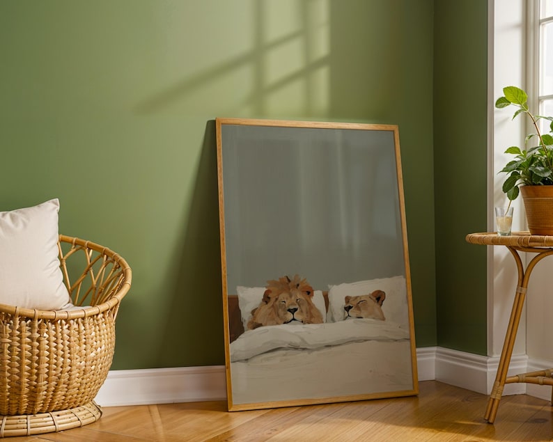 May include: A framed art print featuring a whimsical illustration of two lions in bed, resting on white pillows and under a white blanket. The artwork is set in a room with a green wall and a wooden floor.