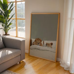 May include: A framed art print depicts a lion and lioness lying in bed. The artwork has a light wood frame and is leaning against a wall in a room with hardwood floors. A gray sofa and a large potted plant are also visible.