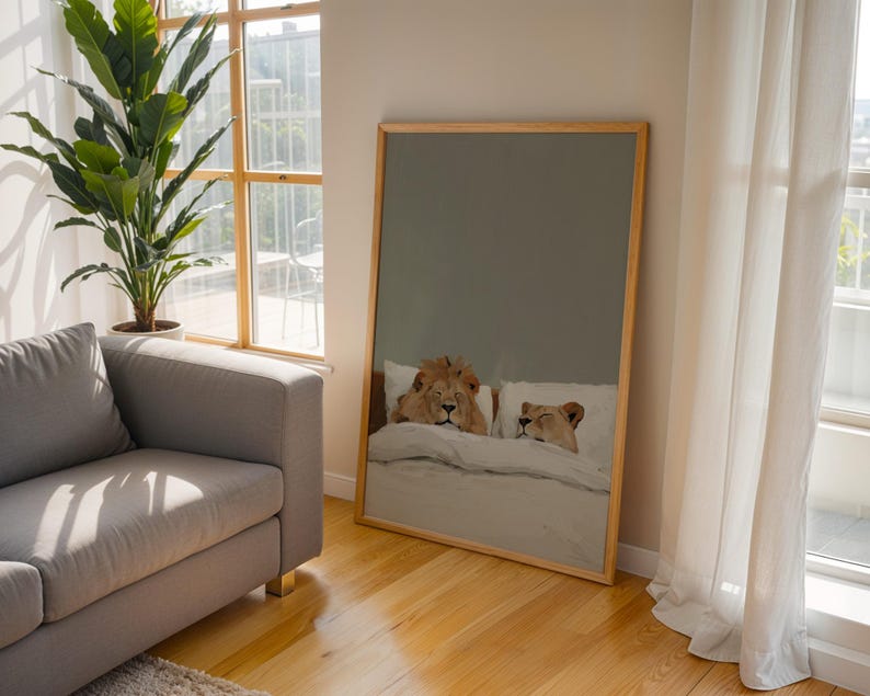 May include: A framed art print depicts a lion and lioness lying in bed. The artwork has a light wood frame and is leaning against a wall in a room with hardwood floors. A gray sofa and a large potted plant are also visible.