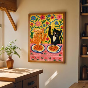 May include: A framed art print featuring two cartoon cats enjoying spaghetti. One cat is orange, the other is black. The background has a floral pattern with yellow, pink, and blue colors. The print is displayed on a wall in a room with wooden accents.