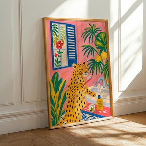 May include: A framed art print depicts a yellow leopard brushing its teeth in a pink bathroom. The illustration includes a blue window, tropical plants, and a sink. The artwork has a gold frame and is leaning against a white wall and wooden floor.