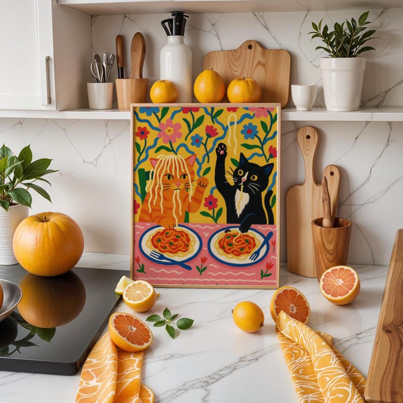May include: A framed art print featuring two cats enjoying spaghetti. One cat is orange, the other is black and white. The background is a colorful floral pattern. The print is displayed on a kitchen counter with lemons, oranges, and wooden utensils.