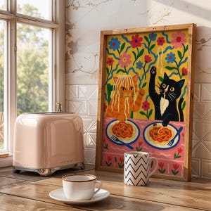 May include: A pink retro-style toaster with chrome accents sits next to a framed art print. The art features two cats eating spaghetti, with a yellow floral background. A white mug with a black zigzag pattern and a teacup are also visible.