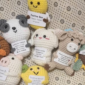 May include: A collection of handmade crochet animals, including a bee, owl, panda, bunny, cow, turtle, pig, and lemon. Each figure holds a small sign with an inspirational message. The figures are in various colors.