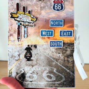 May include: A Route 66 themed scrapbook with a road and motorcycle scene. The cover features signs for a cafe, Route 66, and directional signs. The scrapbook is held by a hand and tied with a white ribbon.